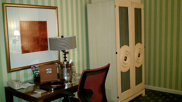 A hotel room desk with a striped lampshade, ice bucket and telephone beside a tall cream armoire and green striped wallpaper