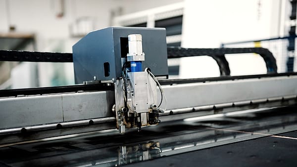 A gray cutting head with a small blue cylinder riding a steel gantry above a dark machine bed in a workshop