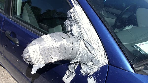 A car's side mirror wrapped in thick layers of silver duct tape against the blue door and dusty window
