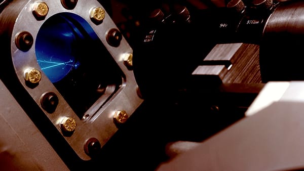 A thin blue beam glowing inside a bolted metal viewport on a dark laboratory vacuum chamber