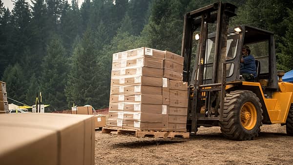 A yellow rough terrain forklift lifting a pallet stacked with cardboard cartons on a dirt clearing