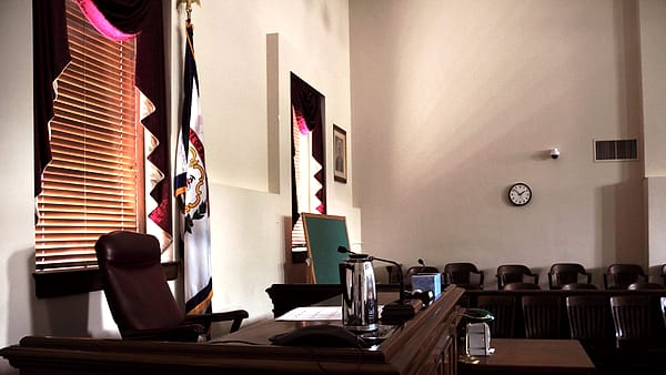 An empty leather chair behind a raised wooden desk in a tall room with a state flag and wall clock