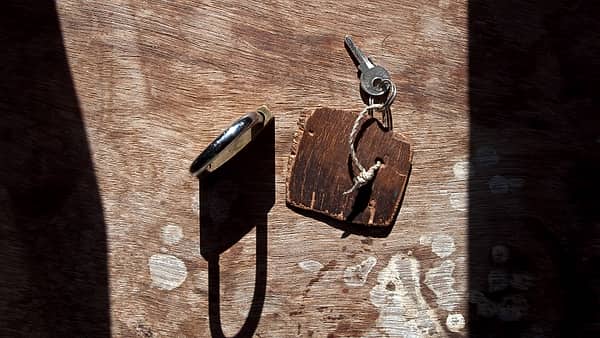 Two keys on a ring tied to a small wooden tag beside a dark metal fitting on sunlit weathered wood