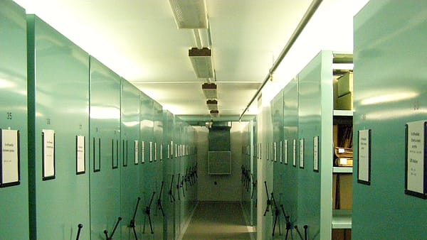 A narrow aisle between tall pale green rolling shelving units with crank handles under fluorescent lights