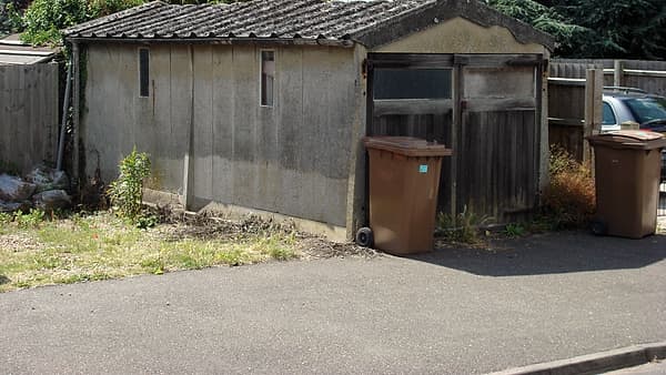 A weathered concrete garage with a tiled roof, battered wooden doors, and two brown wheeled bins beside it