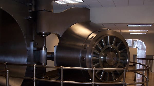 A massive round steel vault door standing open on its hinge behind a low brass rail in a quiet room