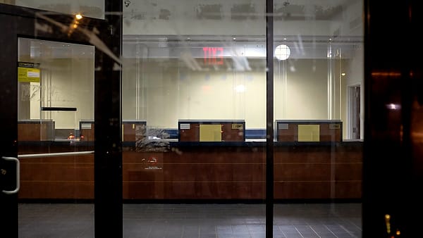 Wood-paneled teller counters inside a dark, empty lobby seen through tall glass doors beneath a lit exit sign