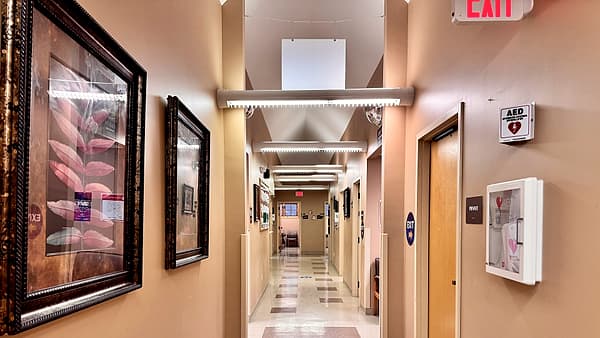 A narrow corridor with framed botanical prints on tan walls and a wall-mounted defibrillator cabinet