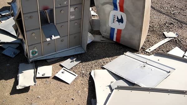 A toppled bank of metal mailboxes and an overturned collection box lying among scattered panels on gravel