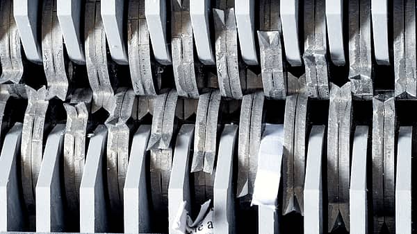 Rows of interlocking steel shredder blades seen up close, with torn white paper strips caught between them