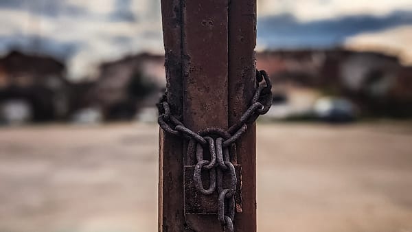 A heavy iron chain wound tight around a rusted metal post, with a blurred empty lot and clouds behind