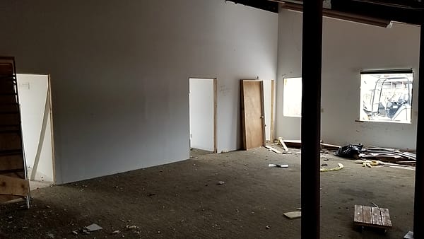 A gutted interior with bare drywall, an unhung wooden door leaning by a doorway and debris on the floor