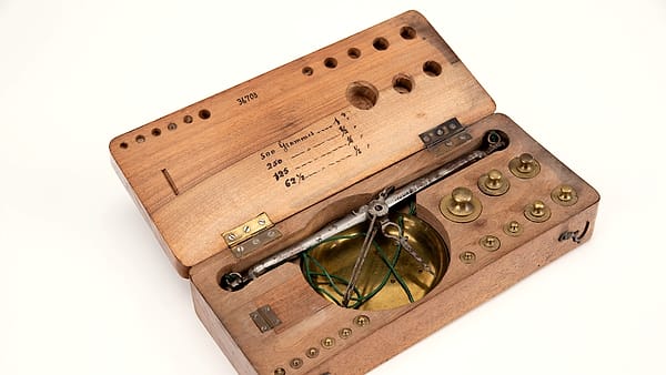 An antique balance in an open wooden case, its brass pan hung on cords beside a row of round weights