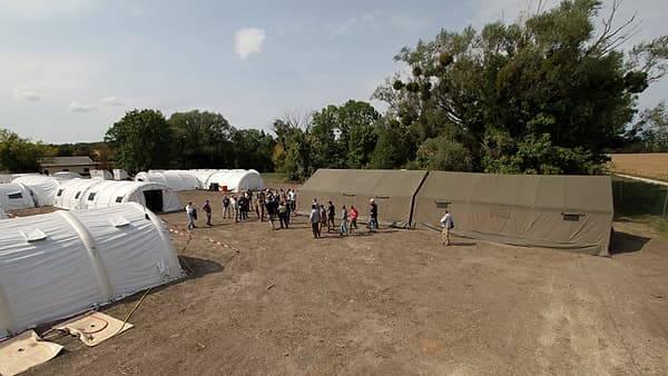 White inflatable arch tents and a large olive-green tent pitched on bare ground, with a small group standing between them