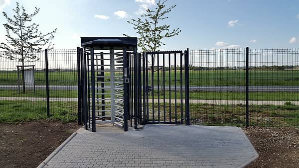 A full-height steel turnstile with a small roof standing in a green mesh fence on a paved apron beside grass