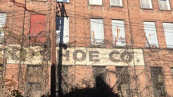 A weathered red brick factory facade with boarded windows, climbing vines and a faded painted sign