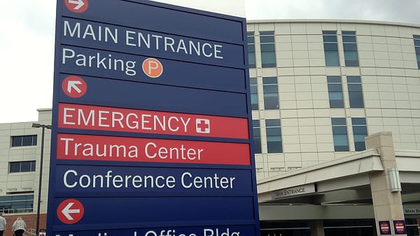 A tall blue and red wayfinding sign with arrows for main entrance, parking and emergency outside a modern building