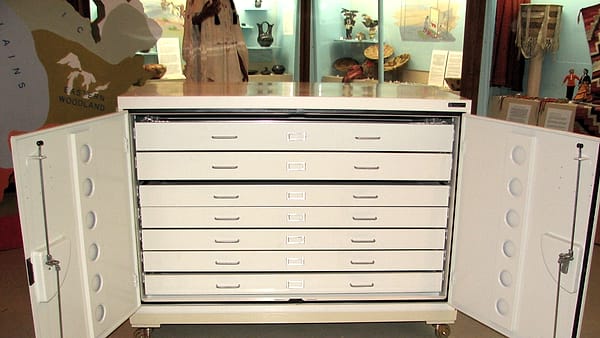 A white metal flat-file cabinet with its side doors swung open, standing in a gallery of glass display cases