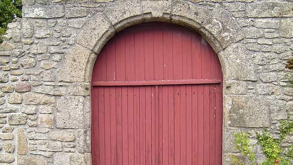 A red painted plank door under a stone arch, set in a rough gray granite wall with ivy at one side