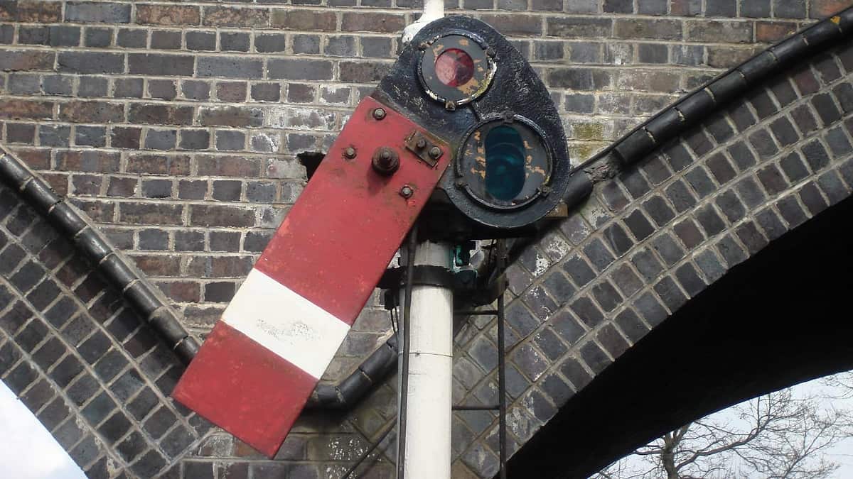 A red and white semaphore signal arm on a white post with its black lamp head against a brick arch