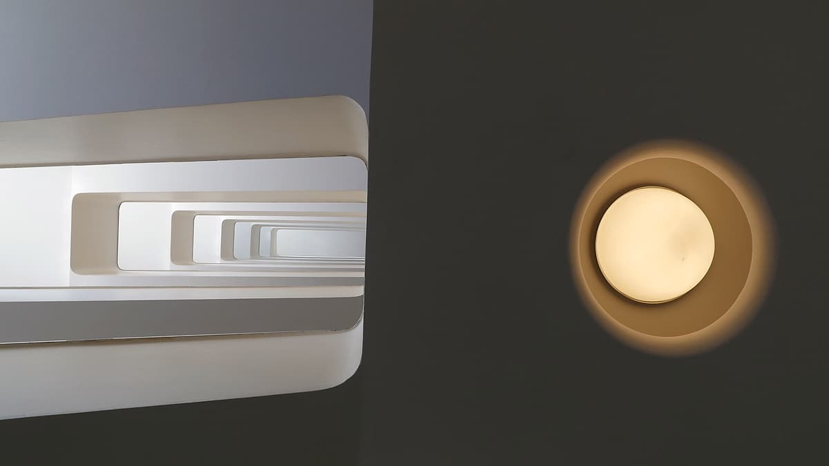 A white rectangular spiral stairwell seen from below, beside a round glowing wall light on a dark wall