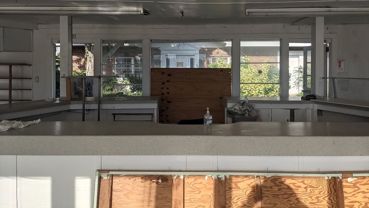 A long laminate counter runs across a stripped room, with bare shelves, plywood sheeting and sunlit windows behind