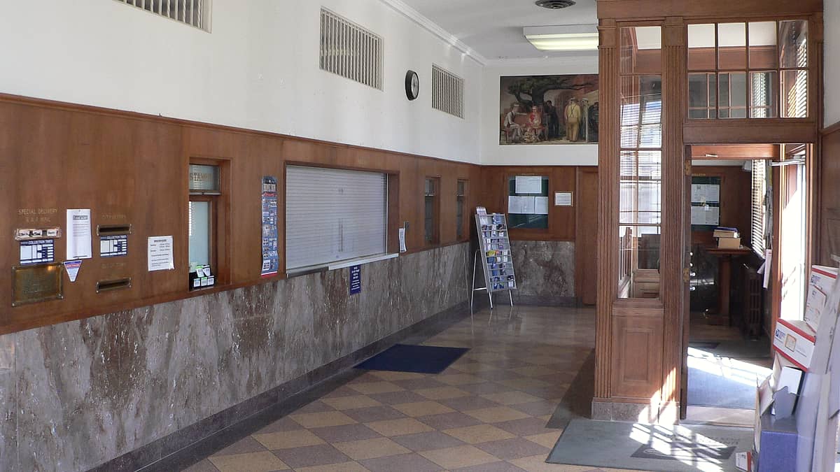 A quiet lobby with a shuttered service window, marble wainscot, wall clock and a checkered tile floor