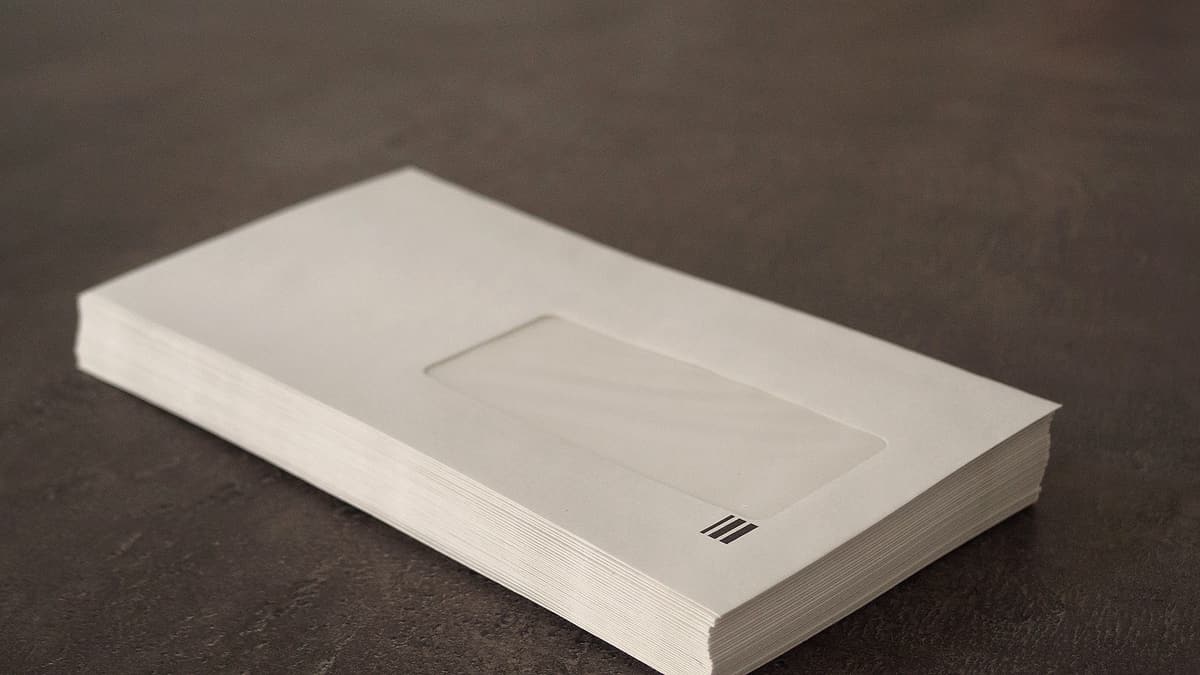 A thick stack of plain cream window envelopes resting on a dark textured tabletop, lit softly from the left